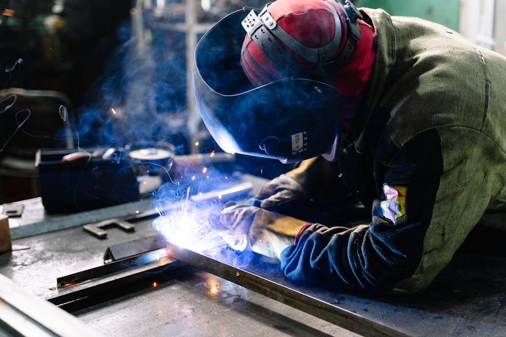 A welder works on metal fabrication indoors, creating bright sparks and smoke.
